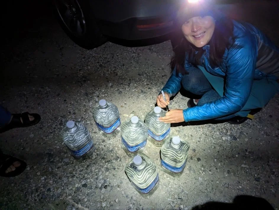 Plastic water gallon jugs with names and dates written in black marker for desert caching.
