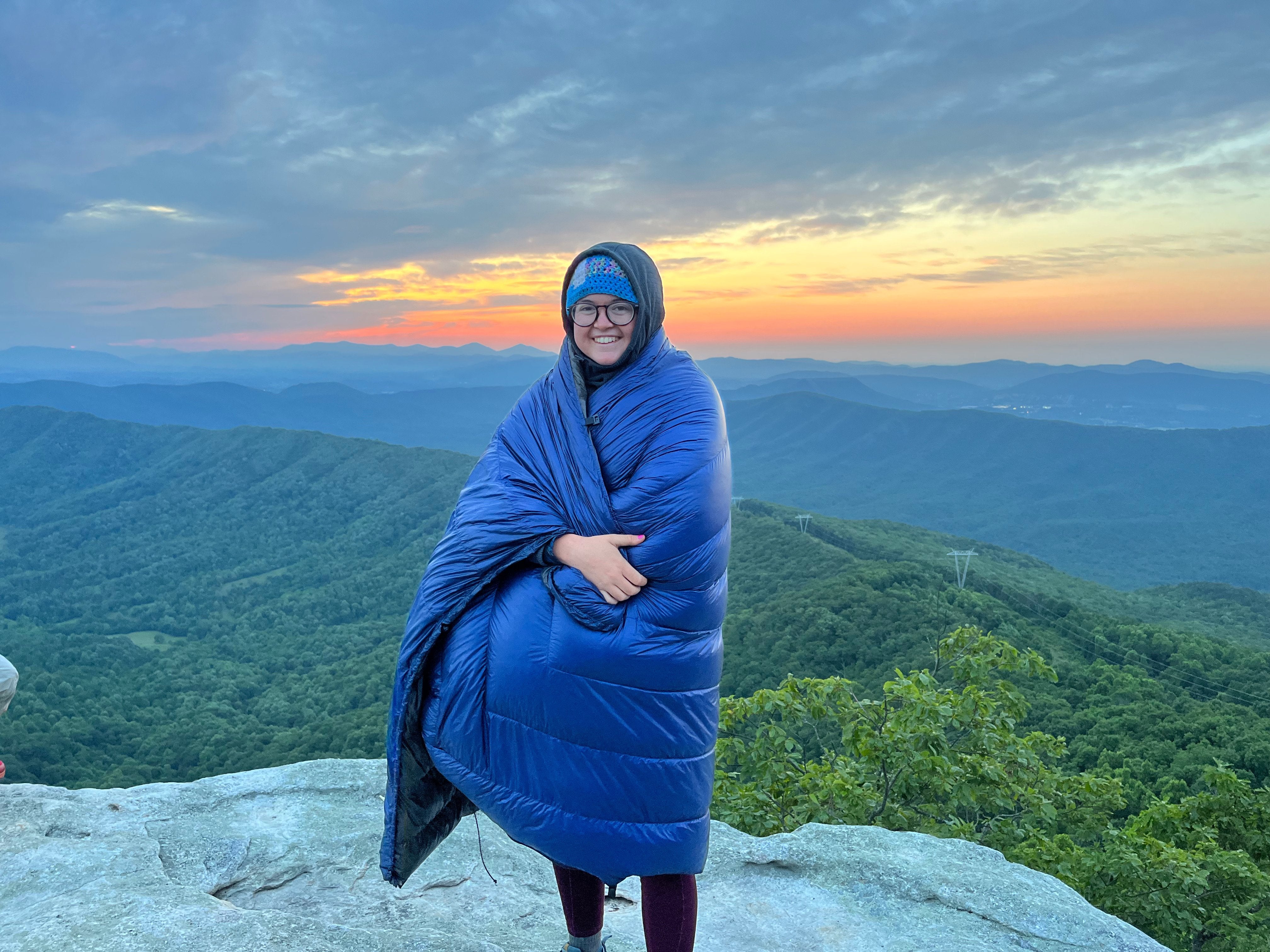 A hiker wrapped in a warm sleeping bag looking out at a mountain sunset.