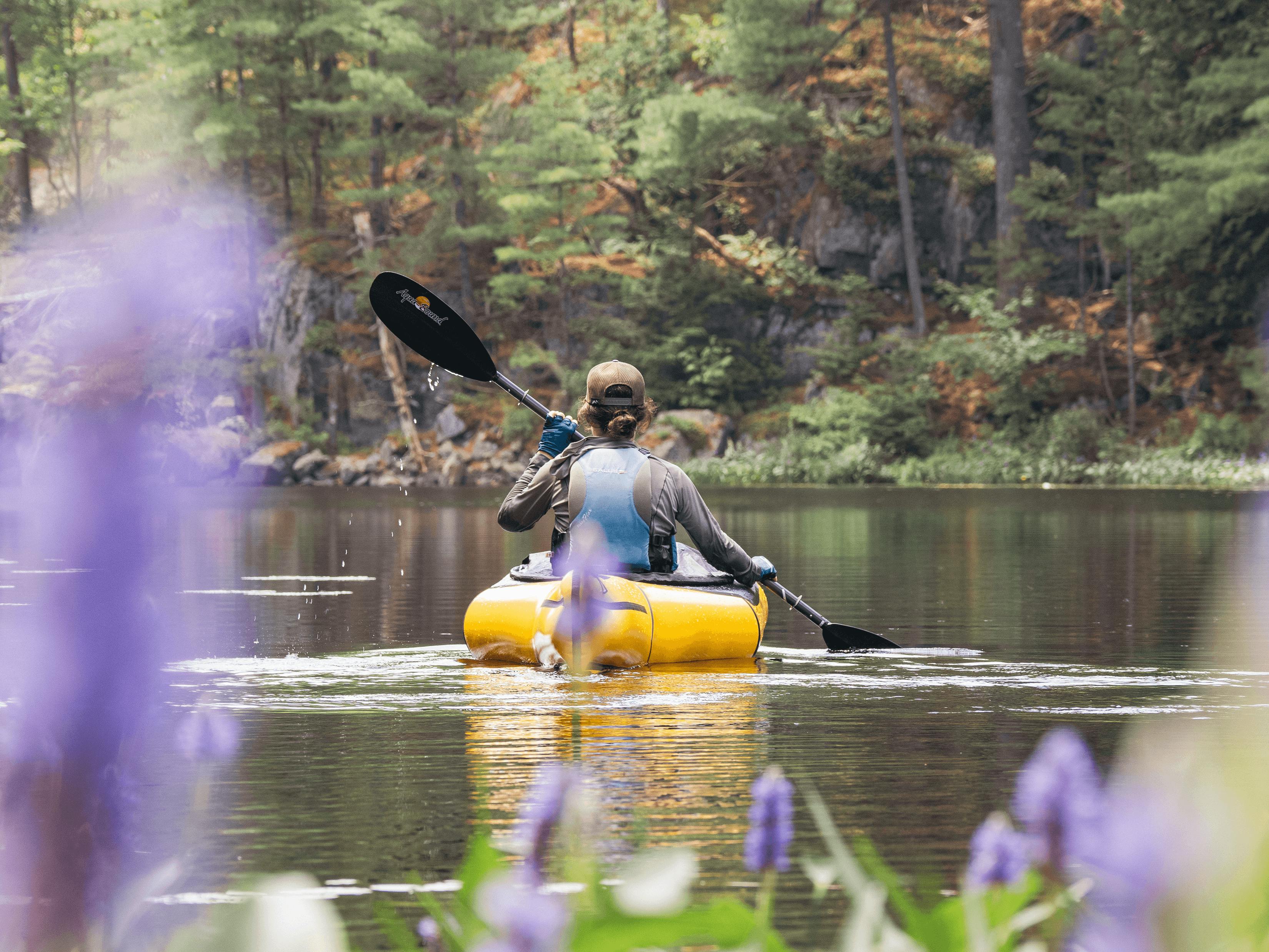 Paddling the Aleutian packraft through a calm coastal area surrounded by purple flowers.