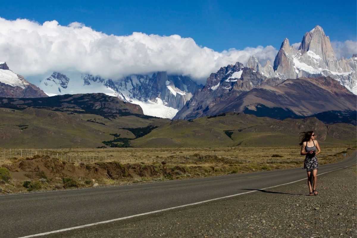 A woman walks down a paved mountain road with high peaks in the distance.