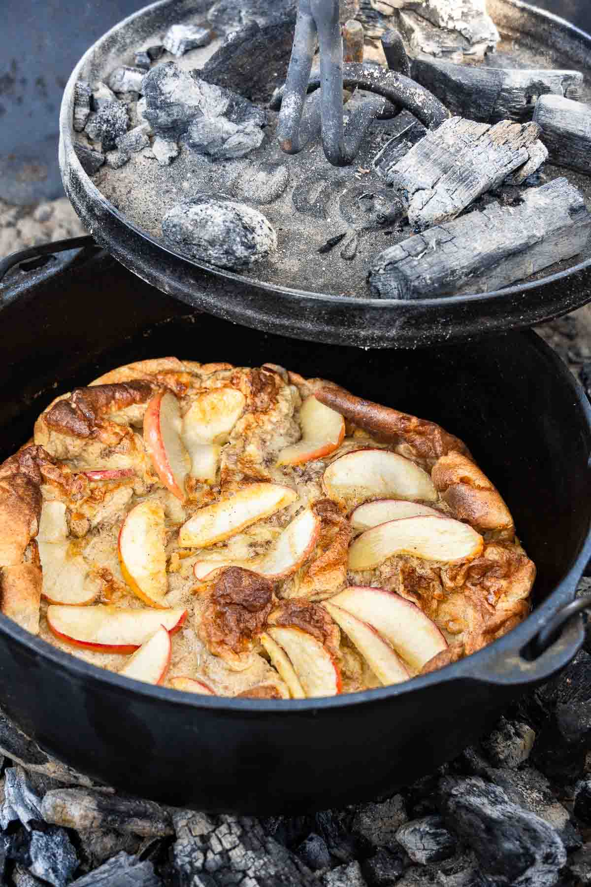 An apple Dutch baby pancake baking in a Dutch oven with hot coals placed on the lid