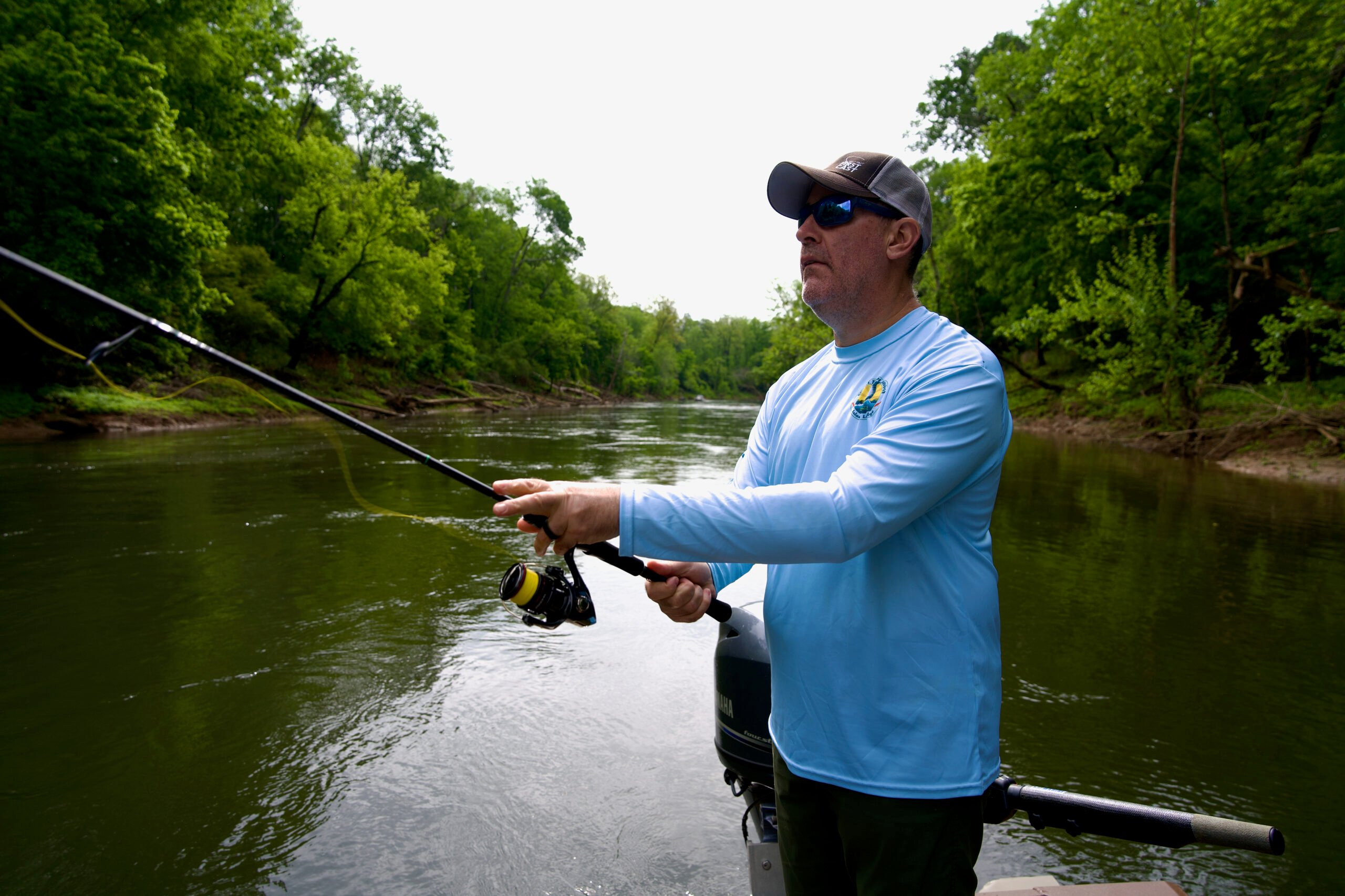 Angler on a boat casting a spinning rod toward a river bank.