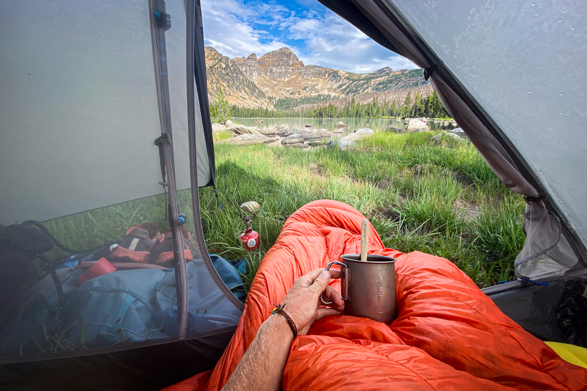 A thru-hiker looks out from their tent at a mountain landscape while wrapped in a gear-focused sleep system.