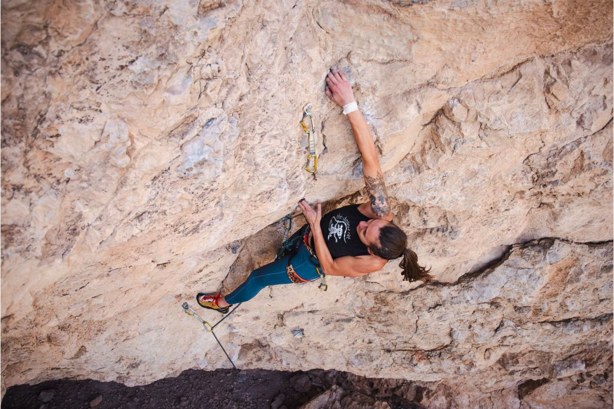 A woman climbs a steep rock face with mountains in the background.